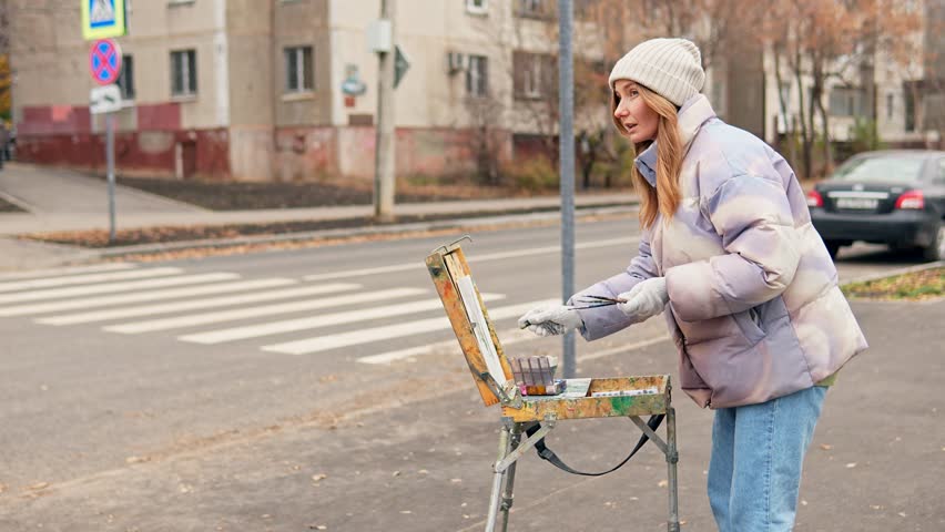 Young adult female artist is drawing with chalk on easel standing on the street at cold autumn day. Caucasian woman is painting urban landscape using portable kit and necessary equipment. Hobby theme.