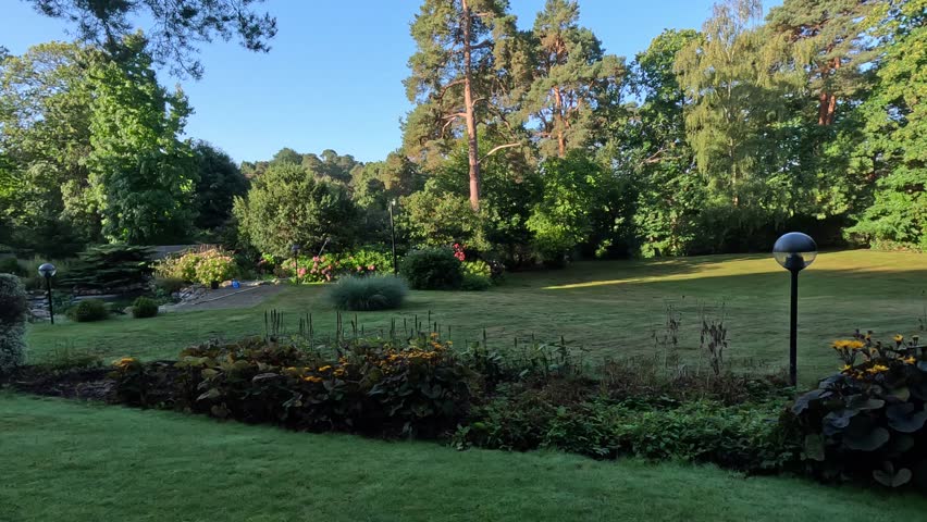 Beautiful garden with green grass, flower beds, and tall trees under clear blue sky on a sunny day. The calm landscape captures peaceful nature and harmony of a well-kept park environment.