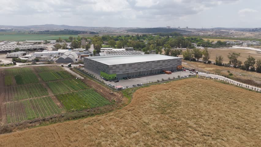 Miluot, Israel - 14 May 2025: Aerial view of a large industrial building contrasts with green fields and dry grass in a landscape under a cloudy sky.