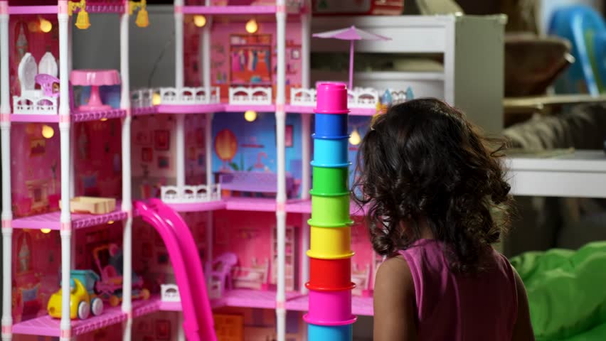 A concentrated little girl sits on the floor, stacking colorful plastic cups in front of a large, brightly lit pink dollhouse with a slide