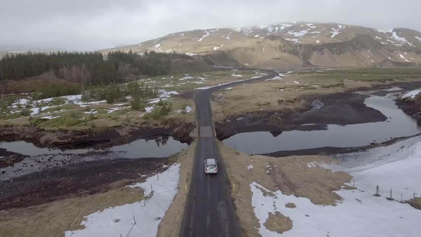 Aerial view of a car driving on a countryside road in Iceland with snow and ice on the side and clouds on background