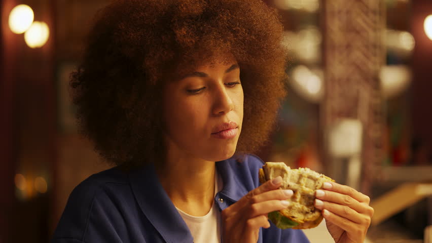 Young black female eating fresh sandwich in restaurant, enjoying tasty lunch