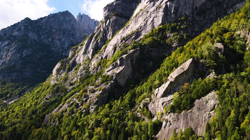 Aerial pullback view of a rugged mountain and boulders covered in pine trees in Val di Mello, Italy. Alpi Retiche, in Val Masino, Valsassina, Italia, flight over torrent with drone. Lake from above.