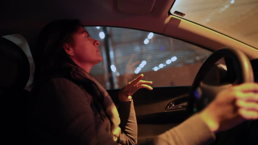 Latina woman driving car at night through city tunnel, one hand on steering wheel, serious expression, light reflections on windshield, motion and urban commute