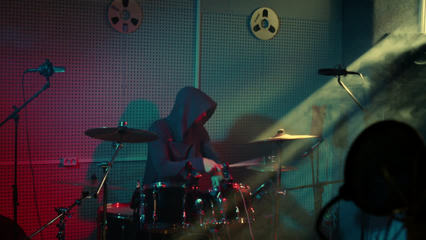 A drummer energetically performs on a drum set, surrounded by vibrant red and blue lighting, creating an immersive and intense musical atmosphere