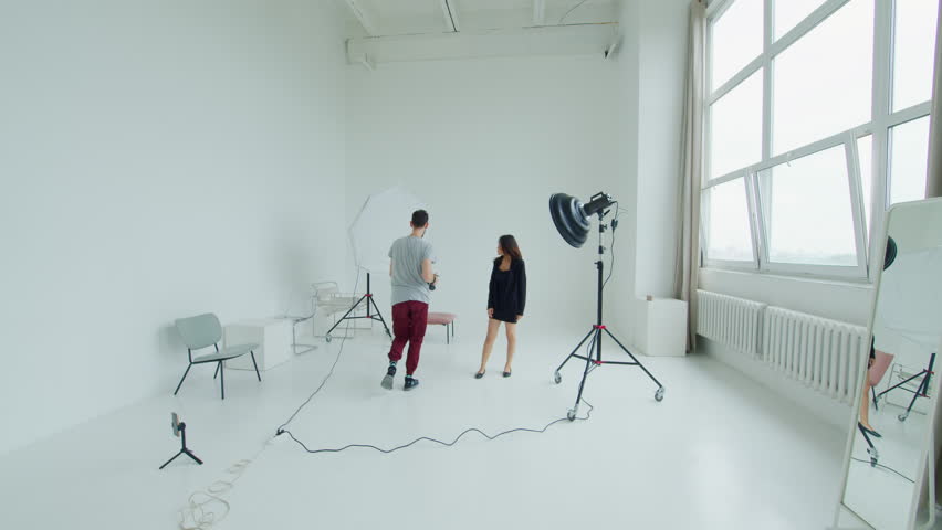 Male photographer shoots beauty portraits of female model in white photostudio. Camera man takes pictures of pretty girl, changes position of soft box. Young woman participates in photography session