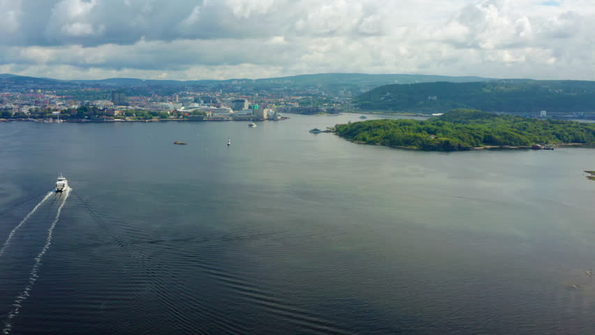Aerial footage of a ferry sailing through Oslofjord with Oslo - Norway capital urban coastline. Summer scenery with green islands, calm sea, and maritime transport. 4K orbit wide establishing shot 