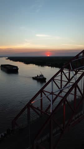 A majestic red bridge spans the wide river and the large barges catch the last golden rays of the stunning sunset. A serene and breathtaking view from above.