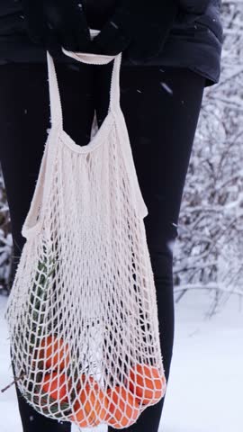 Unrecognizable woman holding Mesh shopping bag with tangerines in snowy winter forest outdoor. Go green concept eco-friendly sustainable Christmas. Responsible