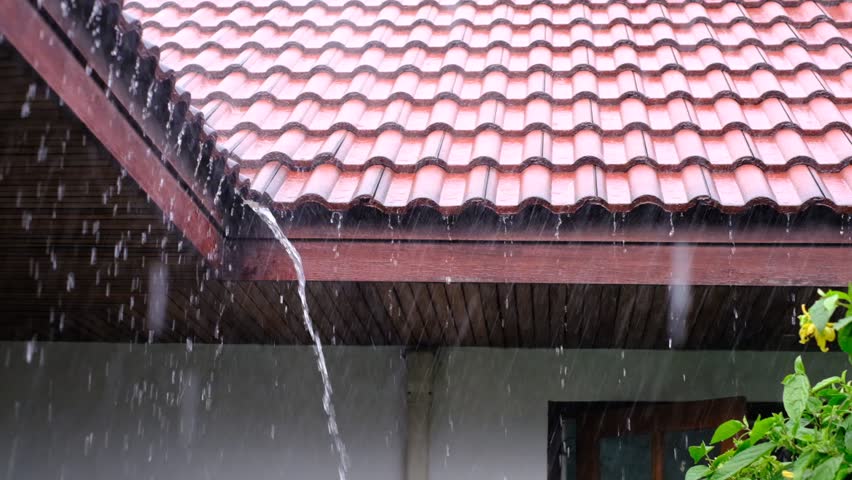 A red roof with rain falling from it. The rain is coming down in a steady stream. The roof is slanted, which allows the water to run off easily