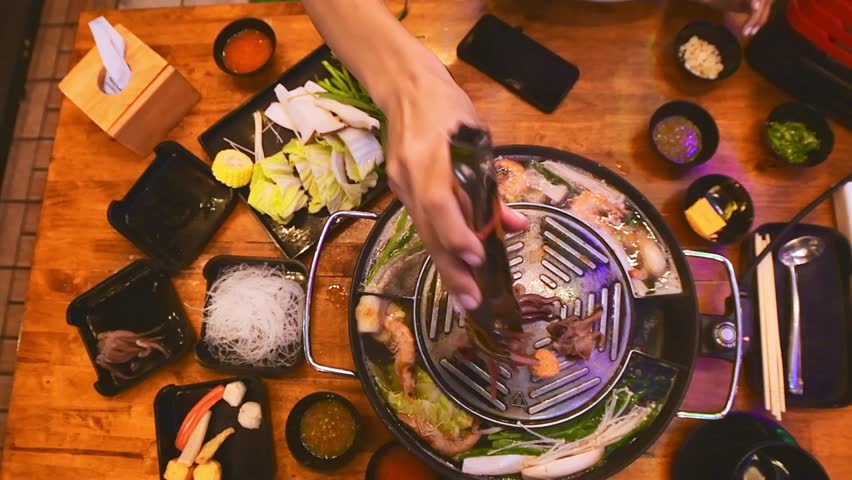 Top view of traditional Thai BBQ hotpot (Moo Krata) with mixed seafood, octopus, shrimp, fish balls, noodles, mushrooms, cabbage, corn, garlic, and dipping sauces on wooden table in Bangkok, Thailand.