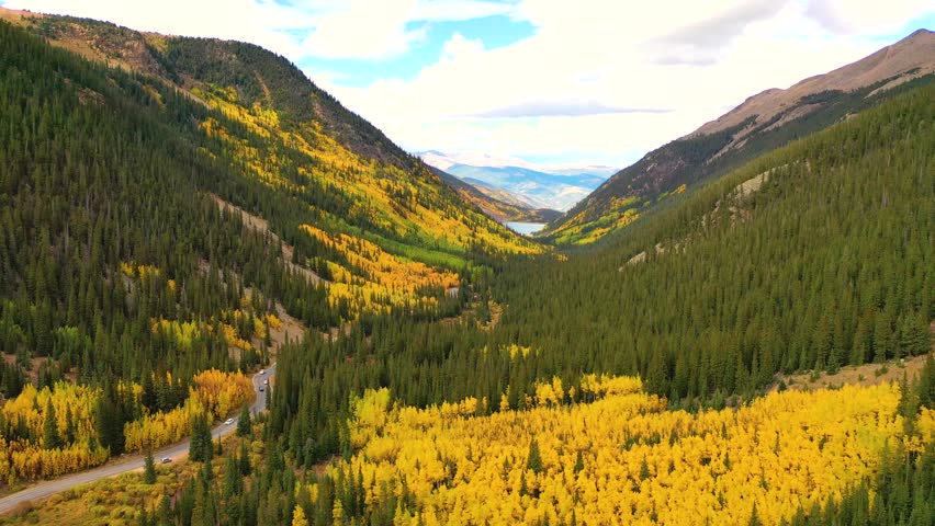 A mountain pass lined with yellow aspens glows under soft sunlight as a few cars travel the twisting road beneath high ridges and calm blue skies.