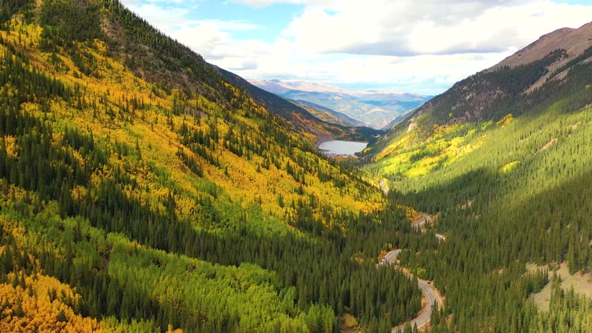 Glacier National Park in Montana bursts with color as forests of gold and green fill deep valleys below snow-covered peaks and winding roads cut through.
