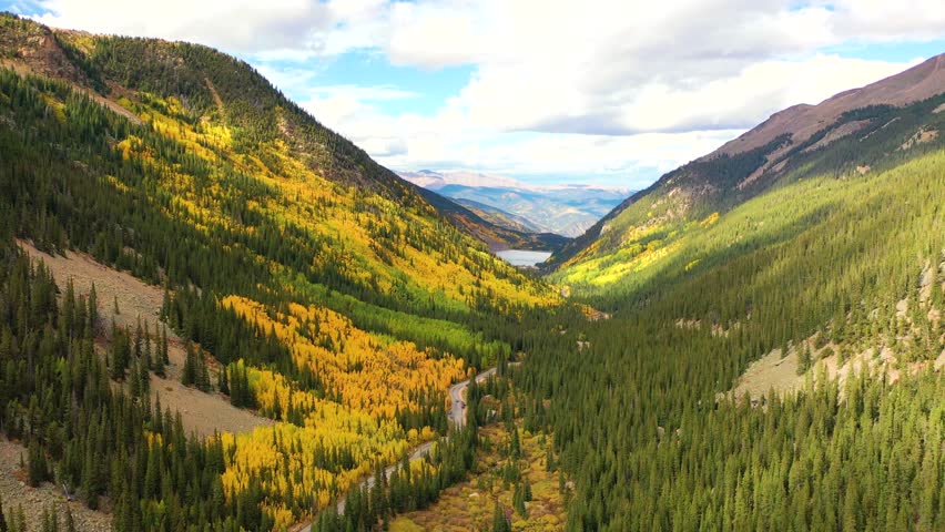 Golden trees blanket the valley floor beneath soft white clouds while ridges of pine and aspen stretch endlessly into the bright blue mountain horizon.