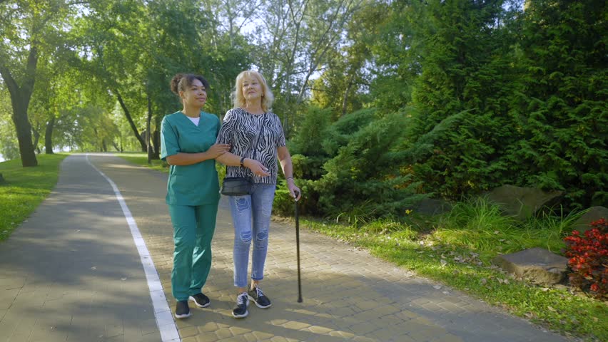 Charming blonde elderly woman using walking cane and positive pretty African American female nurse in uniform taking stroll in summer park, friendly talking and enjoying leisure .