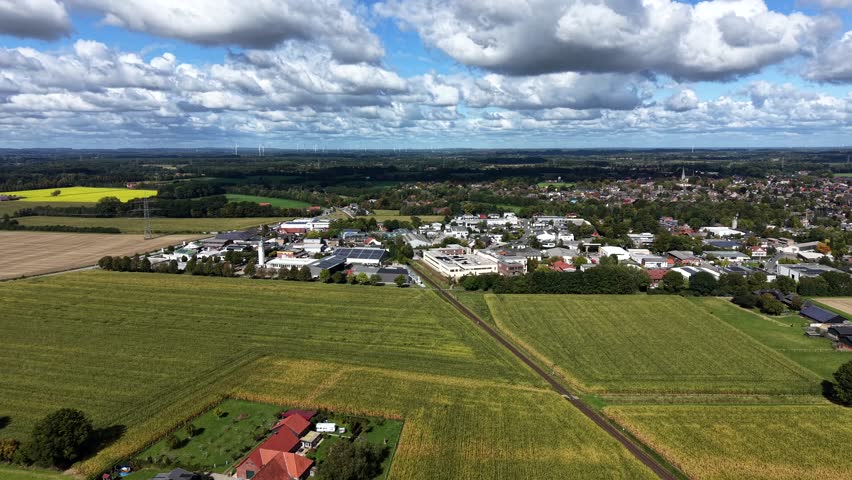 Farm fields in foreground and industrial companies and warehouses in distance. Cloudy summer day in suburb of town. Aerial panorama wide shot.