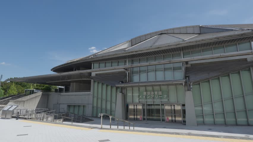 A close-up, eye-level shot of the modern, angular entrance of the Tokyo Metropolitan Gymnasium on a clear, sunny day.