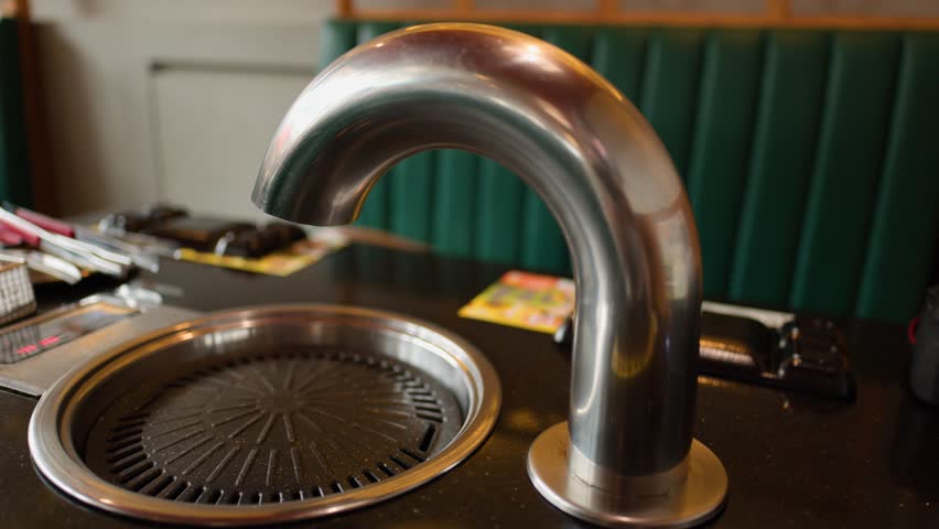 Stainless steel exhaust vent rotates above empty Korean grill pan in warmly lit restaurant booth