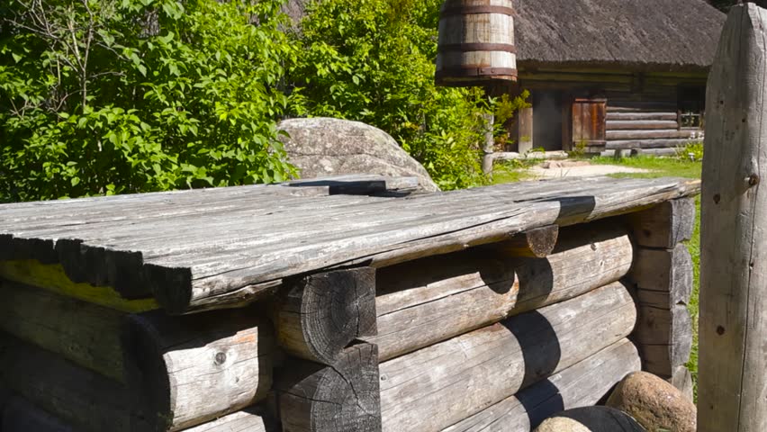 Old traditional wooden brown vintage well with a wooden bucket with rusty straps hanging in front of a old traditional village log cabin with a thatched roof during summer sunny day in a green garden.