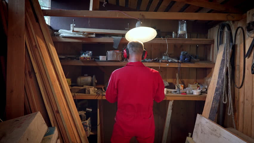 A craftsman stands in front of shelves of parts in his workshop under a swinging lamp.