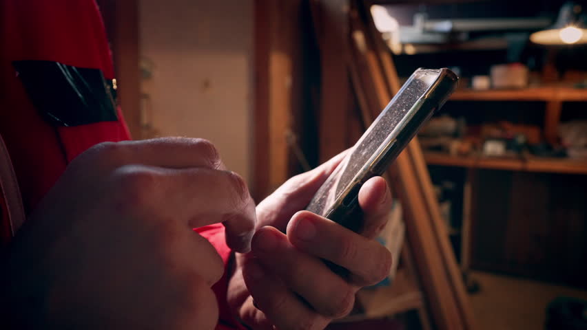 A man in a work uniform is using a smartphone in his workshop, and there is a close-up shot of it.