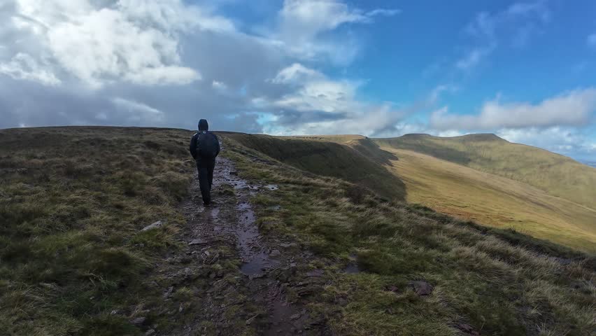 A young man hikes uphill in the Brecon Beacons, Wales. Dramatic clouds, rugged terrain, wide-angle view capture the scenic beauty and outdoor adventure under natural daylight.