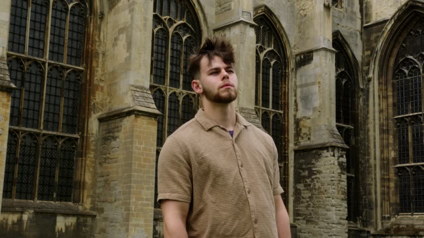 Slow-motion medium shot of a Caucasian Man in casual wear looking around and facing grand Gothic windows of Canterbury Cathedral, England.