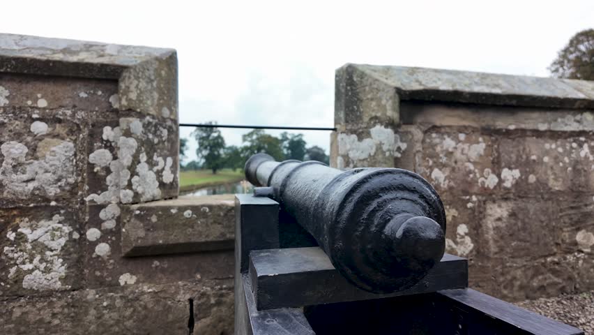 Black iron canon aiming from a stone castle wall towards distant trees and a moat