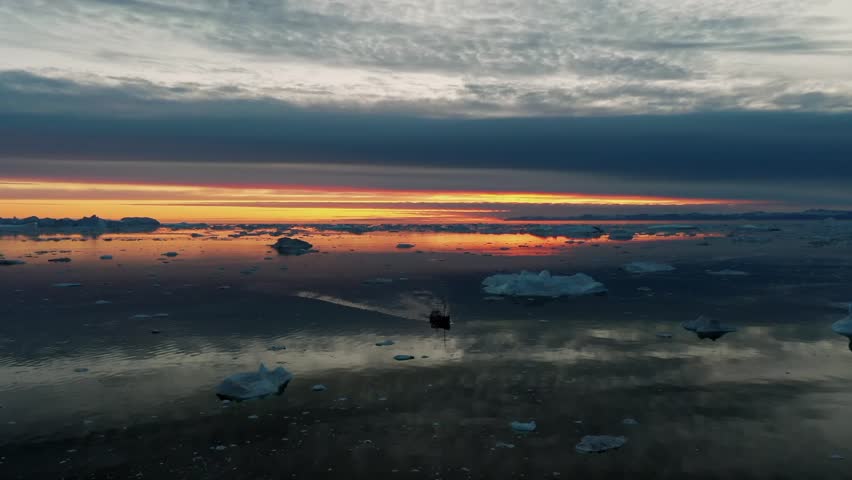 Aerial drone view of a small boat moving through floating icebergs on calm Arctic sea during sunrise in Greenland, with warm orange light reflecting on icy water under dramatic clouds.