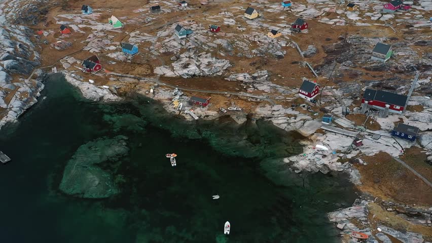 Aerial drone view of colorful wooden houses built along rocky coastline in the remote Arctic village of Ittoqqortoormiit, Greenland, surrounded by clear green water and rugged terrain.