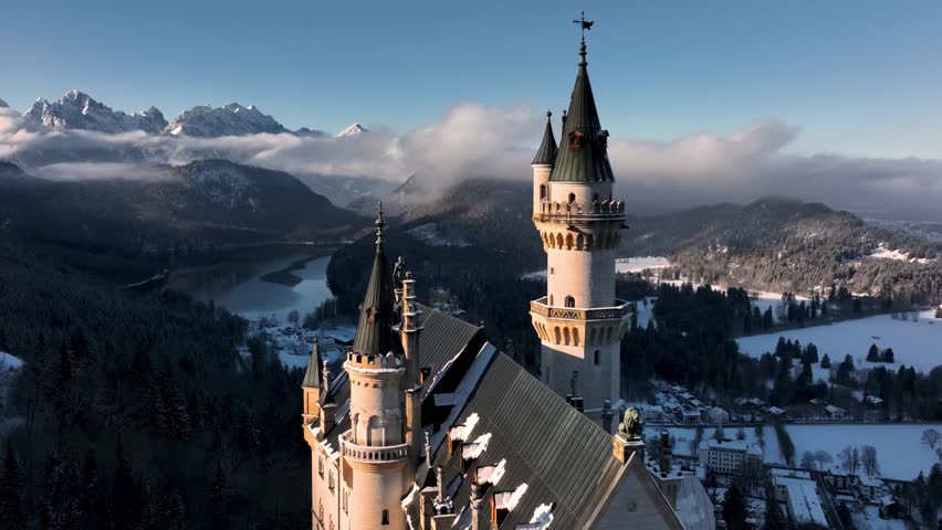 Breathtaking aerial drone view of the fairytale Neuschwanstein Castle in winter. The iconic towers rise above a snowy landscape with a lake and the majestic, cloud-covered Alps behind.