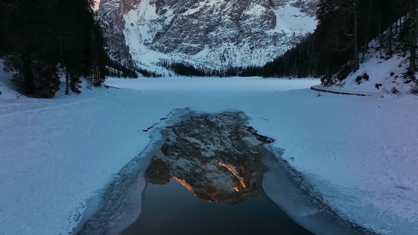 Breathtaking winter view of the frozen and snow-covered Lago di Braies (Pragser Wildsee) in the Dolomites, Italy. A majestic mountain peak, lit by sunrise, is reflected in the water.