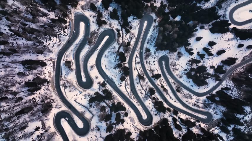 Graphic top-down drone footage of cars navigating a winding serpentine road with multiple hairpin turns up a snow-covered mountain.