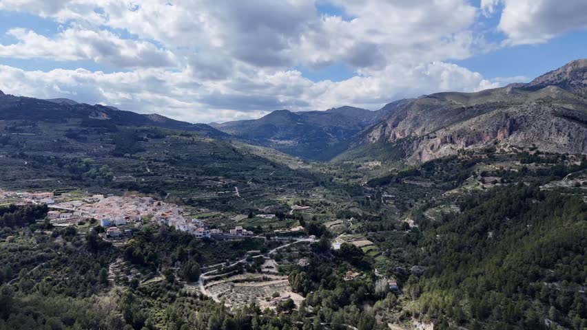 Aerial view of a green valley with terraced fields, small village houses, and surrounding mountain ranges under cloudy blue sky. Scenic Mediterranean landscape.