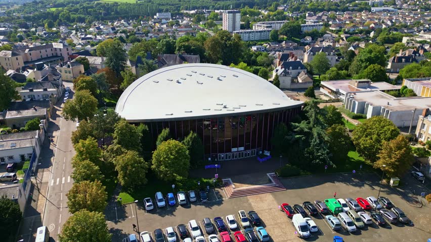 Drone flying backward over Laval multipurpose hall showing parking lot, trees, and city on sunny day.