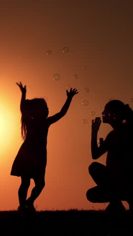 Silhouette of Mother Blowing Soap Bubbles for Daughter at Sunset. Vertical Video