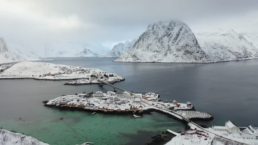 Aerial view of Reine's islands blanketed in snow, a contrast of white roofs against the dark water and snow-covered mountains, Reine, Nordland, Norway.