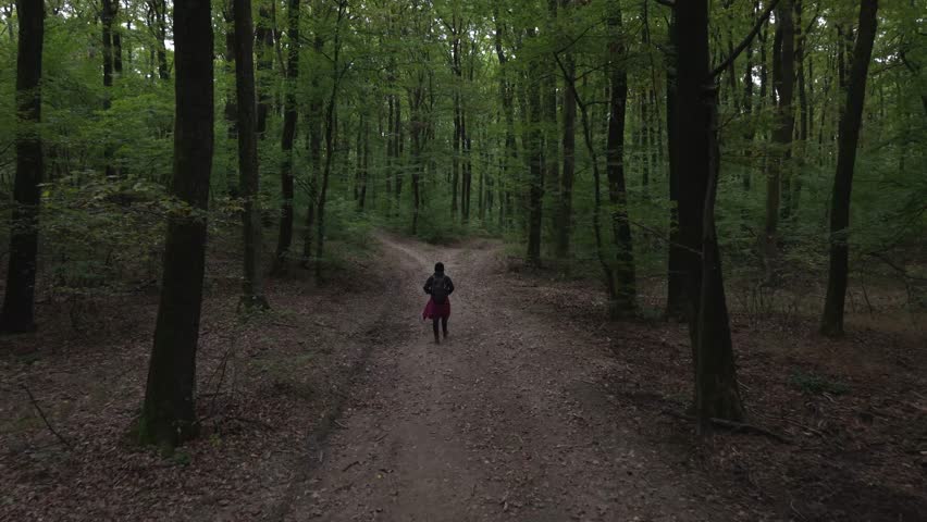 A woman with a backpack walks along a dirt path deep in a dense green forest, approaching a fork in the trail. The peaceful atmosphere and natural surroundings evoke solitude and exploration.