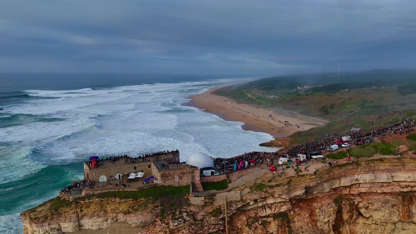 A drone capturing thousands of spectators gathered on a seaside cliff and near the historic fort in Nazare, Portugal, observing huge white waves