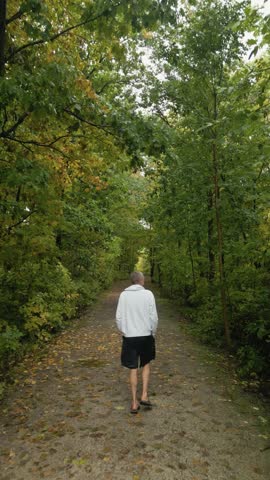 A vertical view of a Caucasian person in a white hoodie walking away on a dirt trail surrounded by trees, creating sense of secluded natural serenity