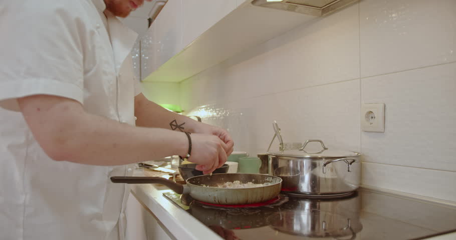 Man cooking in the kitchen with his girlfriend watching him. He is breaking apart pieces of chicken and adding them to a small skillet.