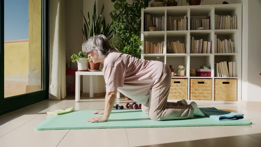 Senior woman with grey hair practicing the cat-cow yoga pose on a green mat in her sunlit living room, arching her back for flexibility, health, and wellness during her morning routine