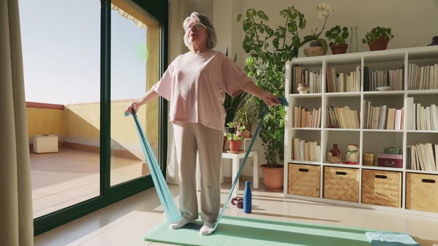 Elderly woman with grey hair performing a strength training exercise with an elastic band, standing on a yoga mat in a sunlit living room and focusing on her physical wellness