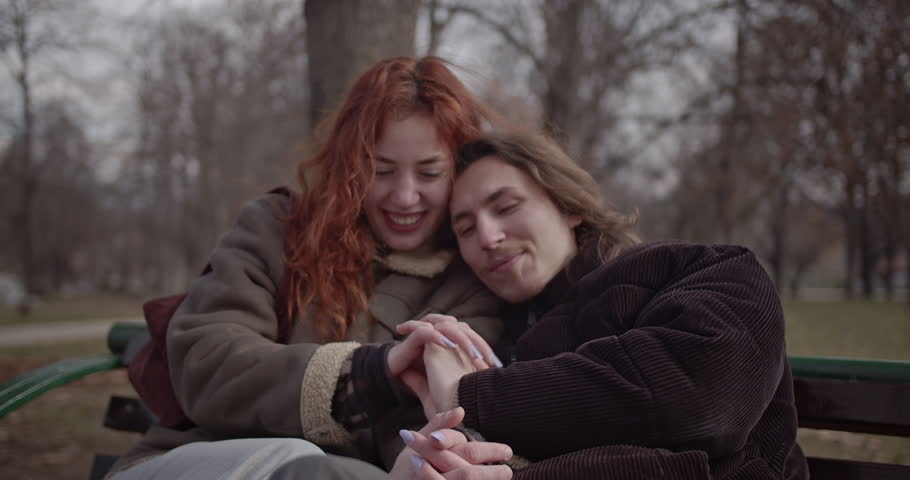 Couple in love sitting on park bench holding hands and cuddling on gloomy autumn or winter day. They look happy, content, and affectionate.