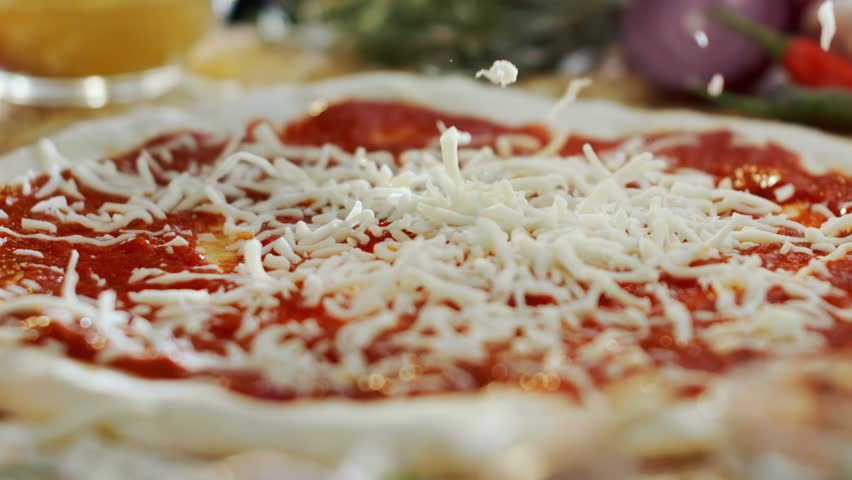 Close-up slow-motion shot of shredded mozzarella cheese falling onto a raw pizza base with fresh tomato sauce, showing the preparation of a traditional italian dish in a restaurant