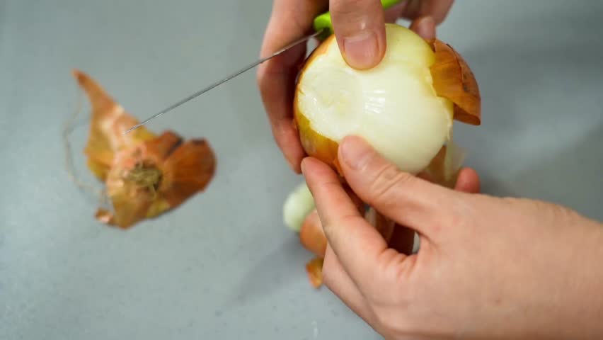 Peeling onion skin with knife and hand above grey cutting board