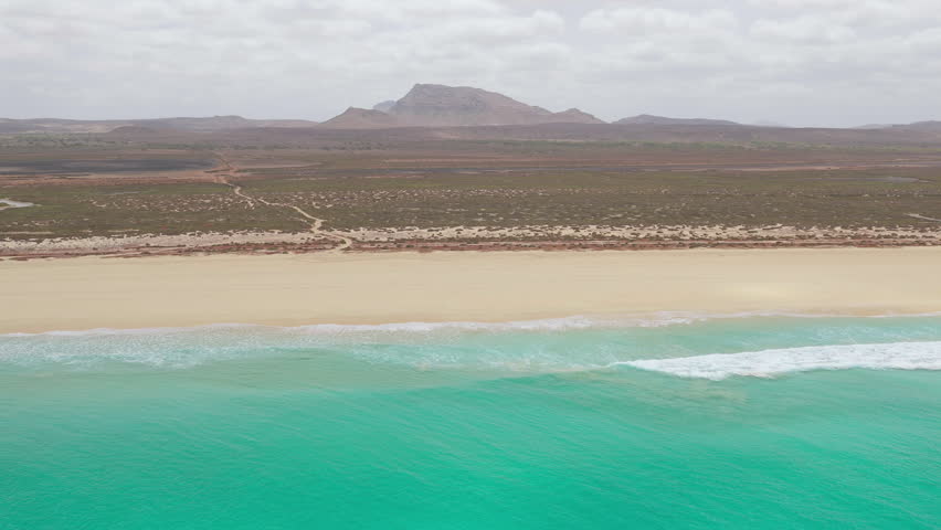 Drone footage of Santa Monica Beach,showcasing endless white sand, turquoise waves, and a pristine,background volcanic mountain and desert,Boa Vista, Cape Verde.