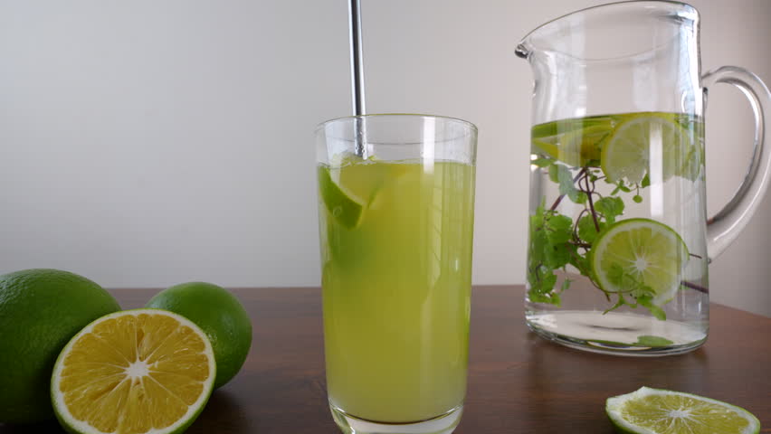 A close-up shot of the fresh limeade in a glass, stirring with a straw and a pitcher with limes and mint