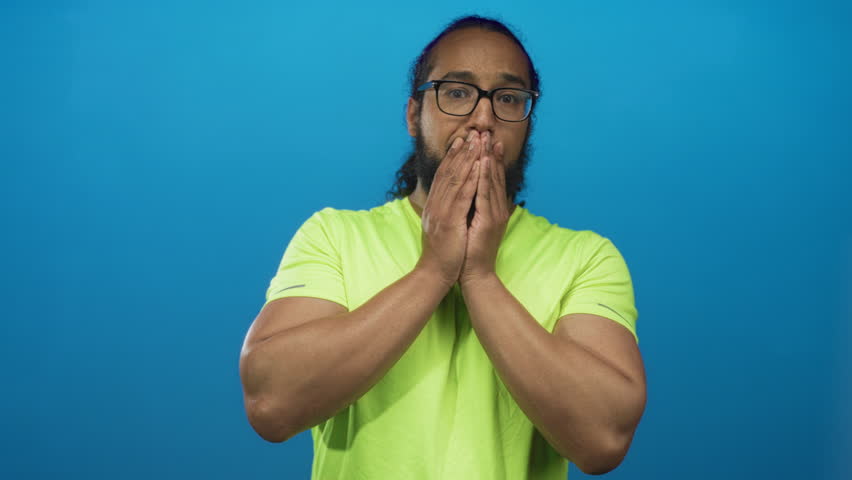 Man with open mouth and clasped hands wearing neon green shirt and glasses in blue studio; surprise reaction.