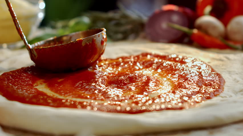 Close up of a professional chef spreading fresh tomato sauce with a ladle on a raw pizza dough, the first step in preparing a delicious homemade traditional italian margherita